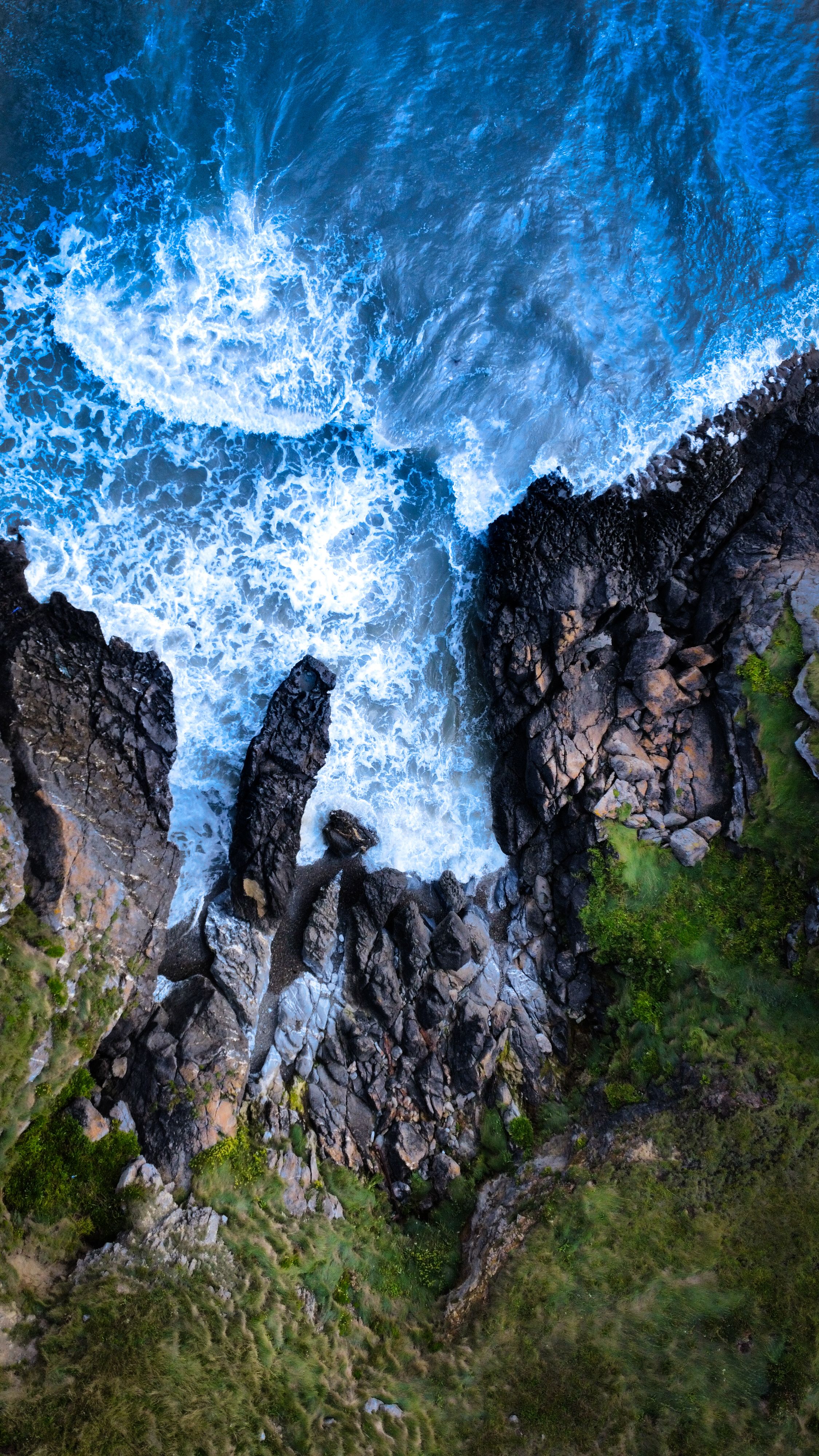 A Breathtaking Aerial View Of Waves Crashing Against Rugged Cliffs And Rocks, Showcasing Nature's Raw Power.