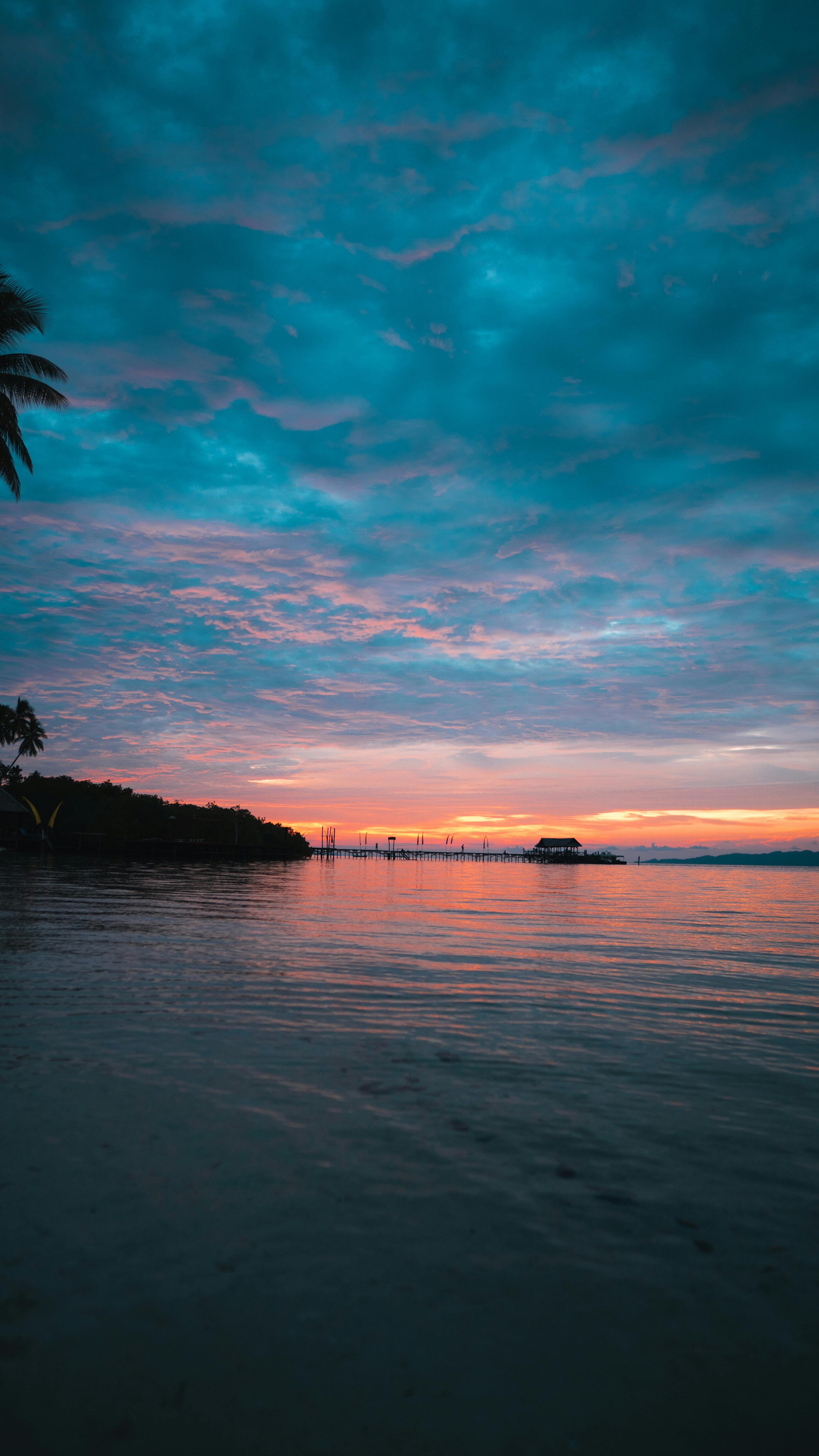Dramatic Sunset Reflecting On The Calm Ocean At Raja Ampat Islands, Indonesia, With Palm Silhouettes.
