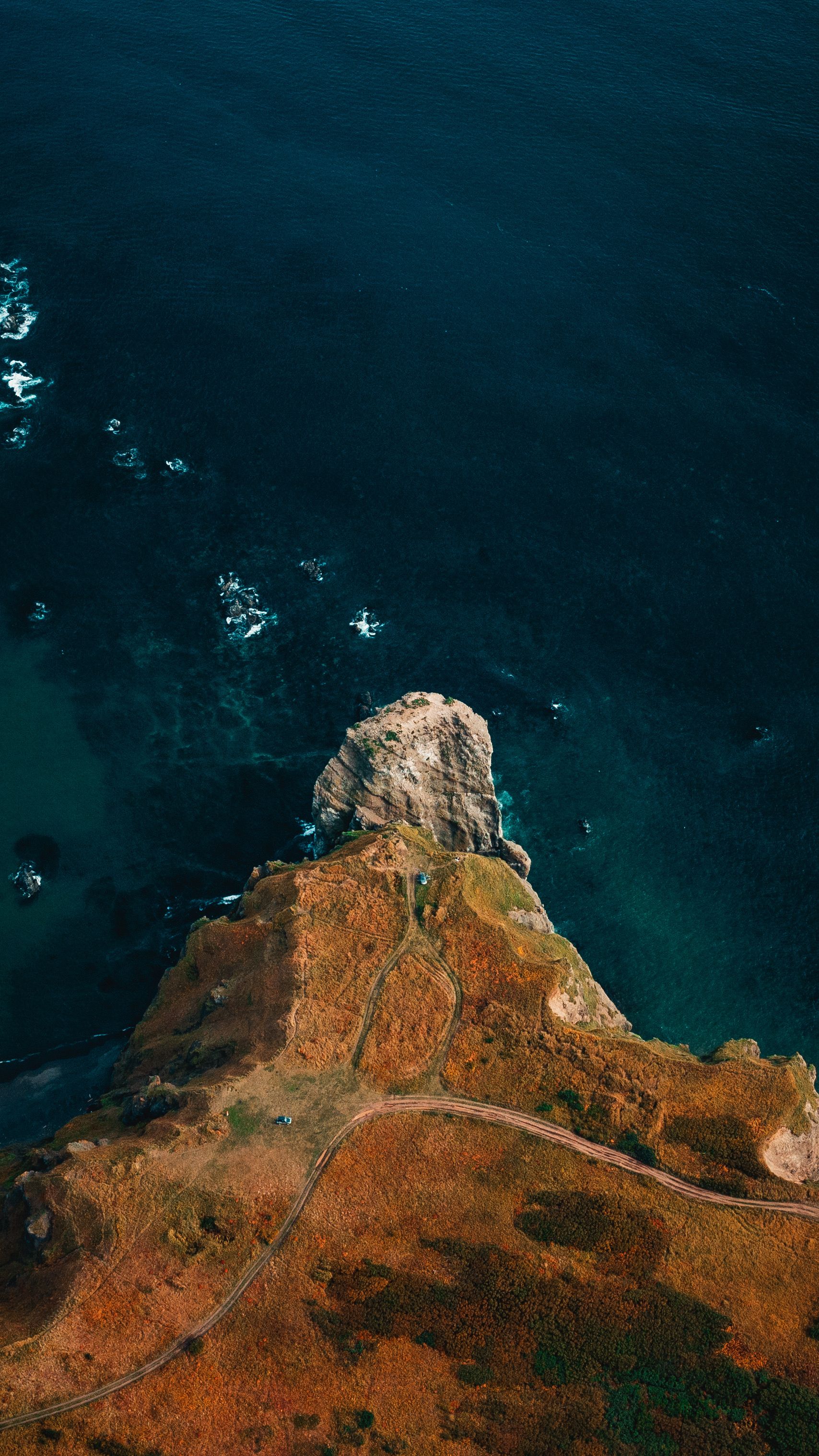A Stunning Aerial View Of A Rugged Coastal Cliff Meeting The Deep Blue Ocean Under Bright Daylight.