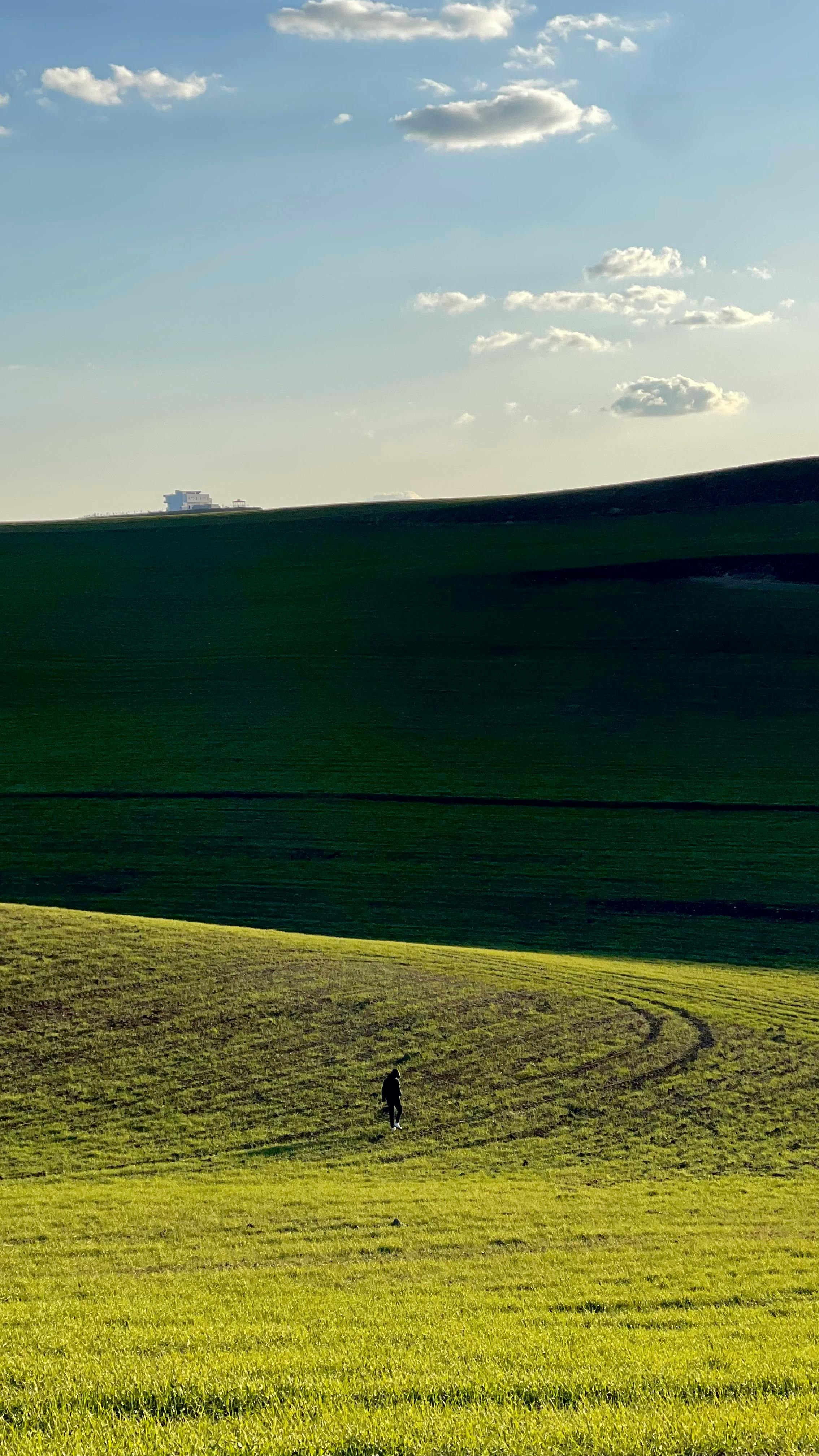 A Solitary Figure Walks Through Lush Green Hills Under A Clear Blue Sky.