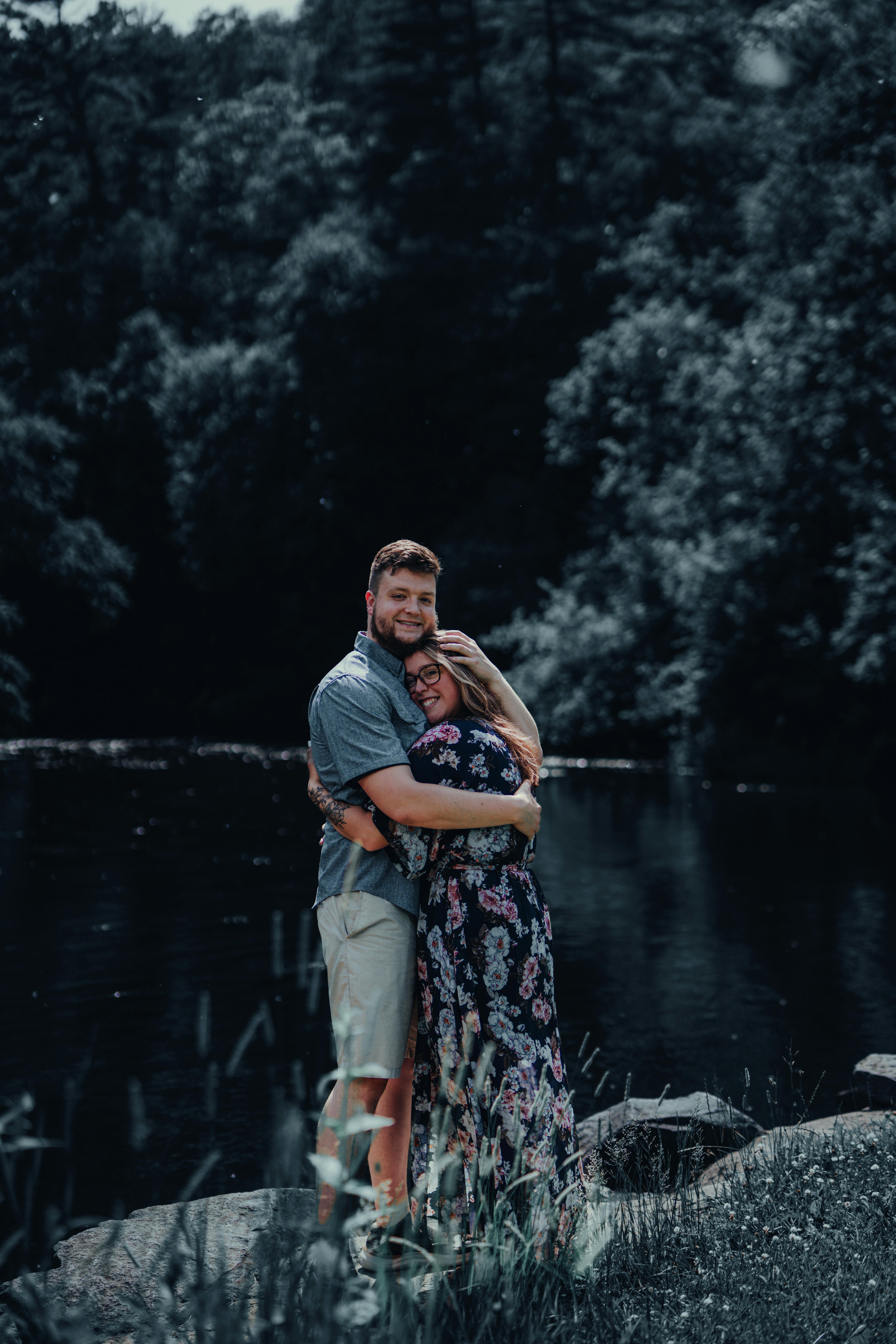 A Loving Couple Embraces By A Peaceful River, Surrounded By Lush Greenery. Perfect For Romantic Themes.