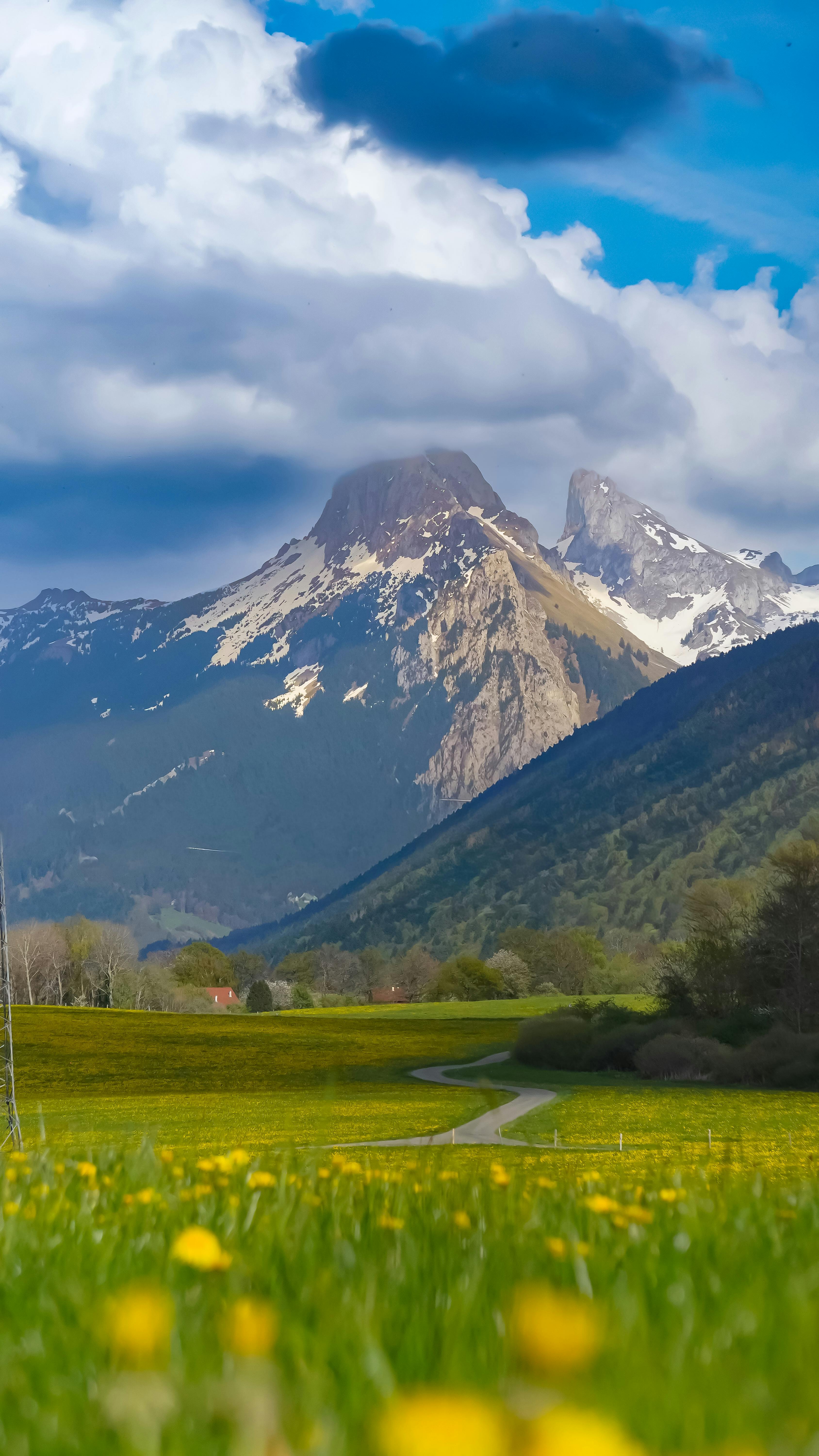 Breathtaking View Of Mountains And A Lush Green Meadow With Fluffy Clouds Overhead.