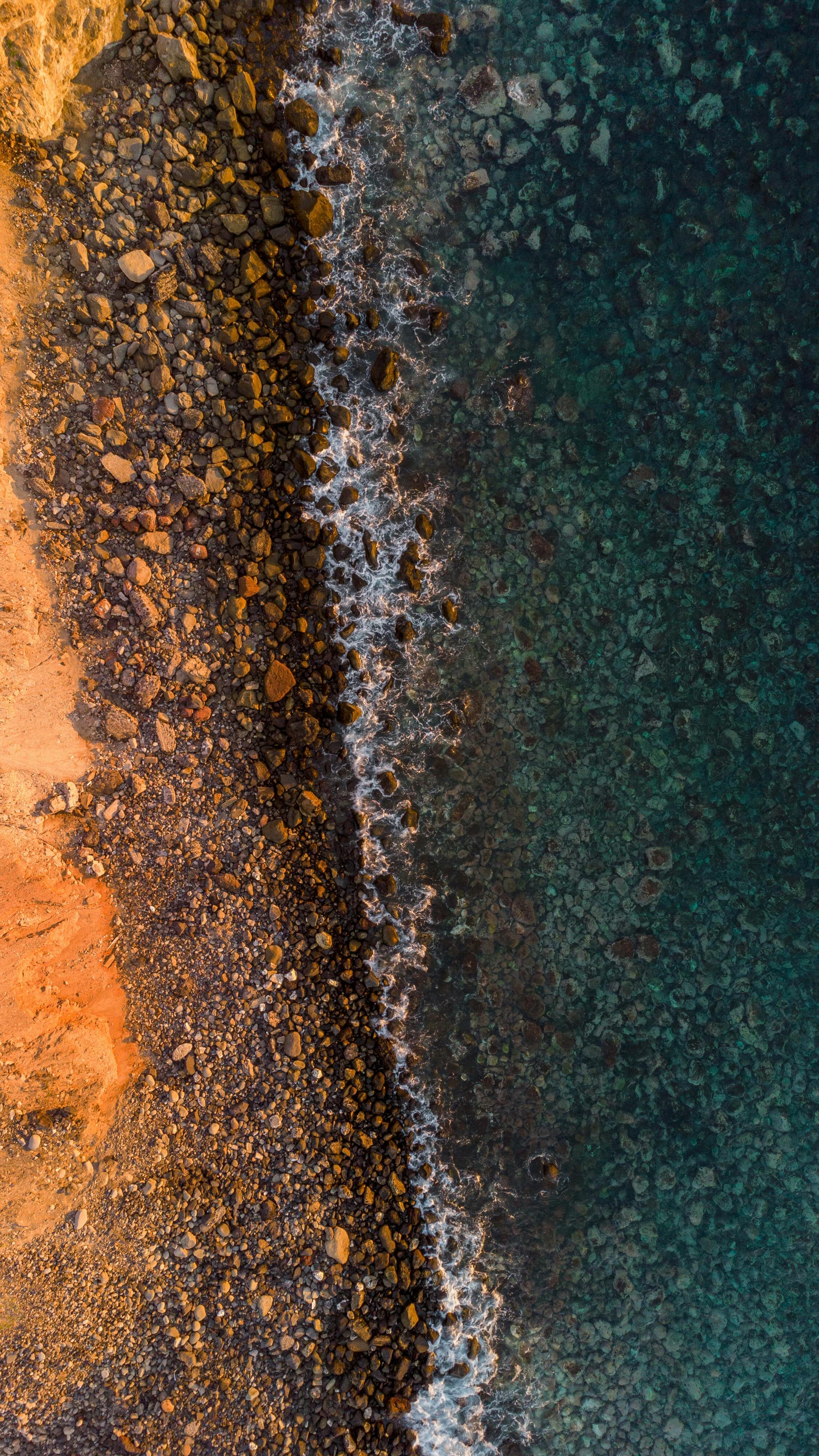 Aerial Shot Capturing The Vibrant Contrast Of A Rocky Coastline Meeting Clear Ocean Waters.