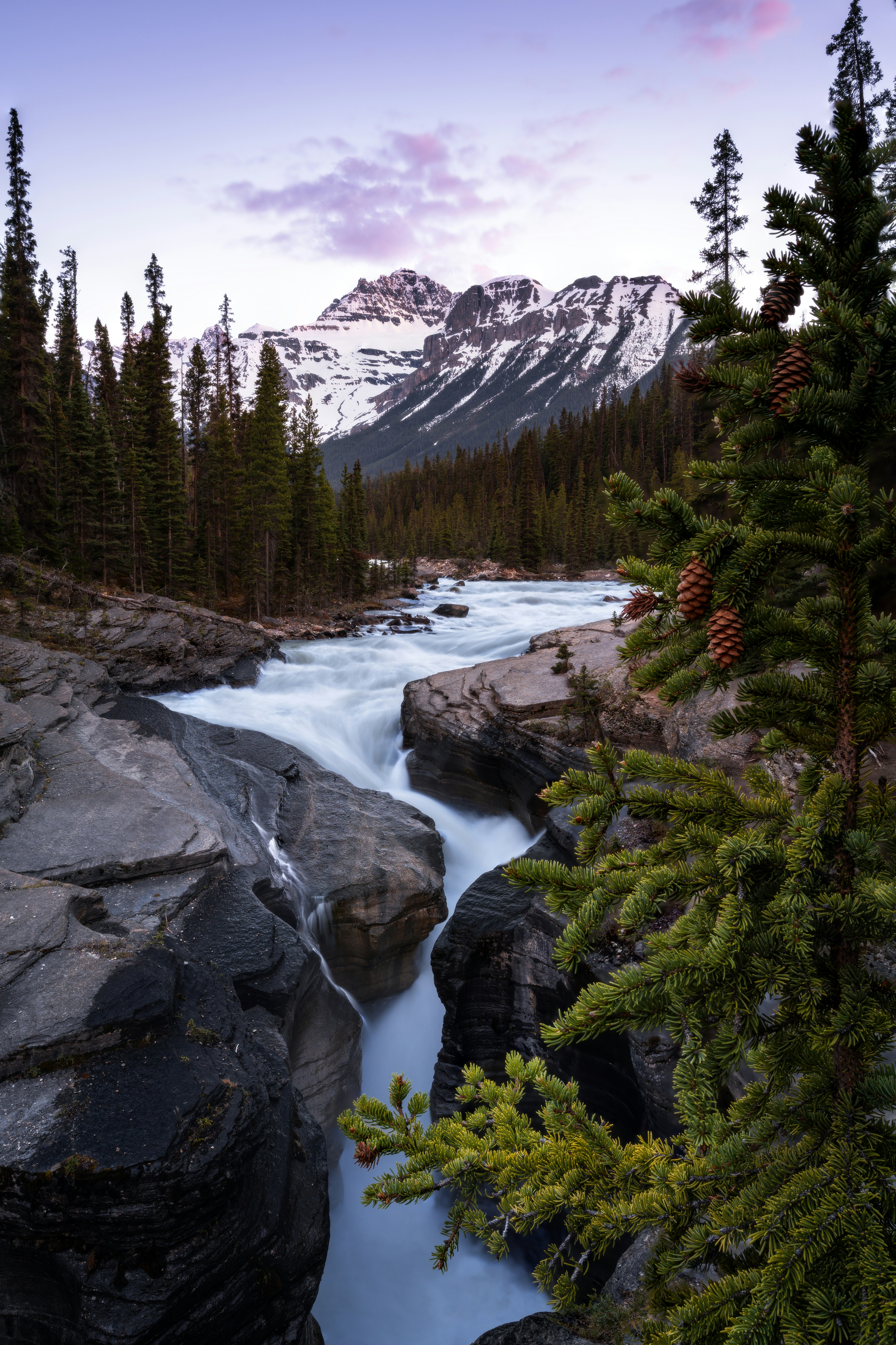 Serene River Cascades Through A Forest With Snow-capped Peaks.