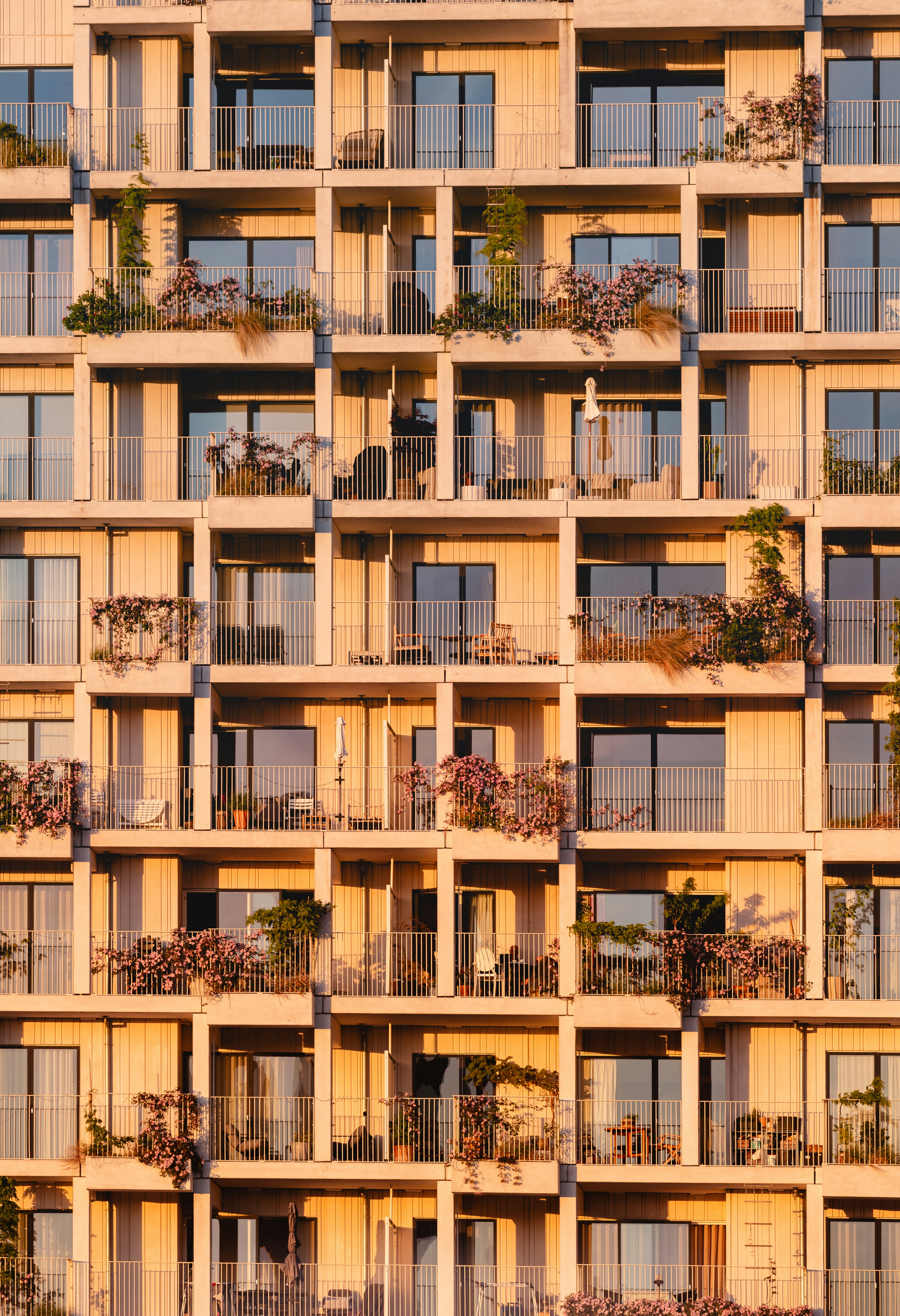 Close-up Facade Of A Modern Residential Apartment Building Glowing In Warm Golden Sunset Light, Showcasing A Repeating Grid Of Private Balconies Filled With Lush Plants, Flowers, And Greenery.
