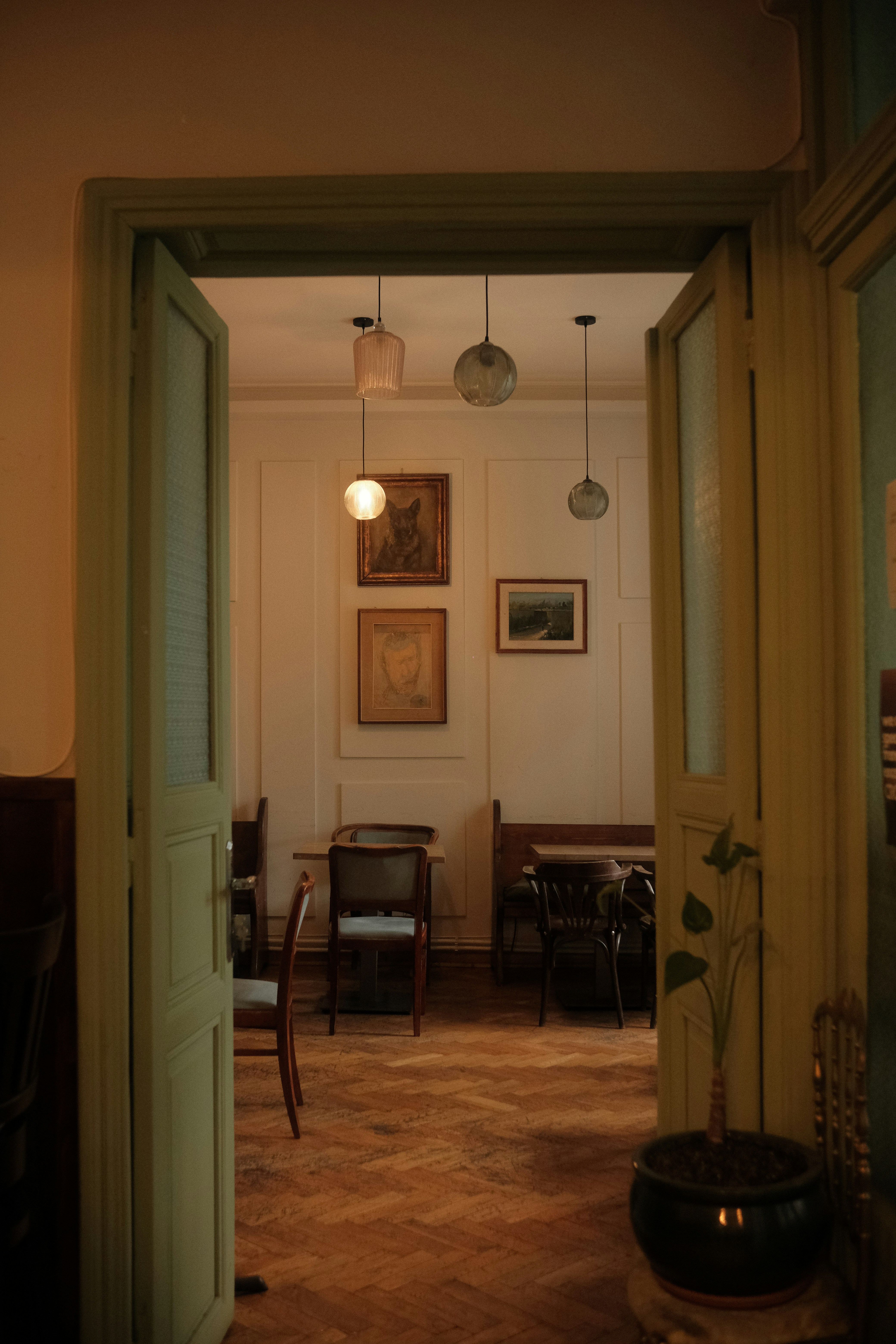 A Softly Lit Vintage Interior Seen Through Open Doors, With Wooden Floors, Hanging Pendant Lights, Framed Artwork, And A Calm Old-world Café Atmosphere.
