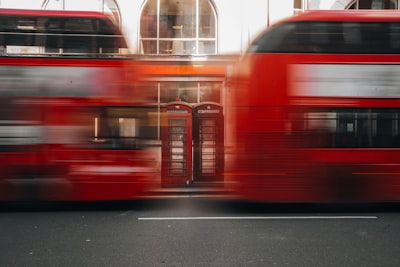 Two Blurred Red Double-decker Buses Passing A Building. wallpaper preview