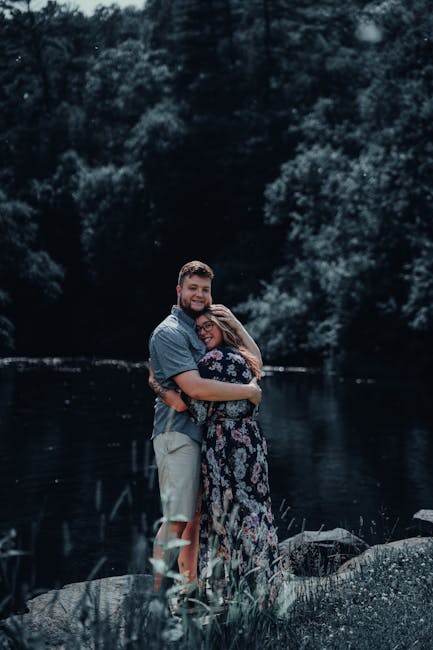 A Loving Couple Embraces By A Peaceful River, Surrounded By Lush Greenery. Perfect For Romantic Themes. wallpaper preview