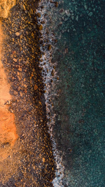 Aerial Shot Capturing The Vibrant Contrast Of A Rocky Coastline Meeting Clear Ocean Waters. wallpaper preview