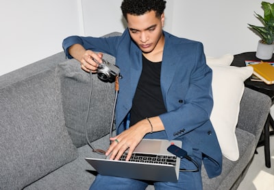 Young Man Using Laptop And Camera On Sofa.