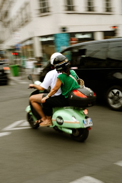 Two People Ride A Green Scooter On A City Street.