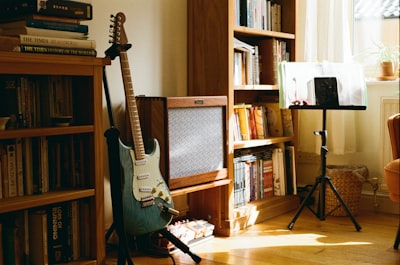 A Blue Electric Guitar Rests Against A Vintage Amp In A Sunlit Room Of Books.