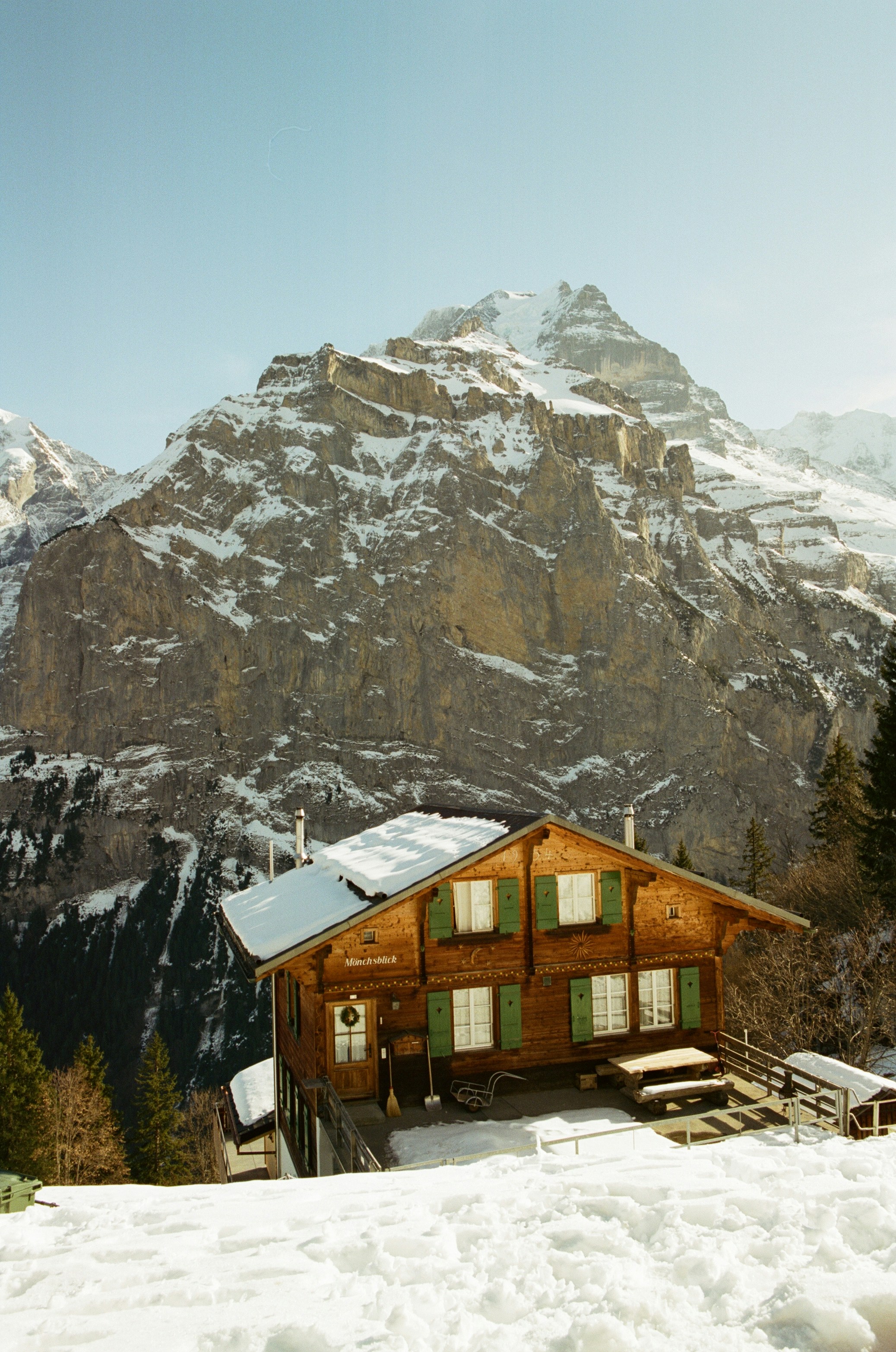 A Timber Mountain Hut With Green Shutters Set Against A Vast Snow-streaked Alpine Peak. HD mobile wallpaper