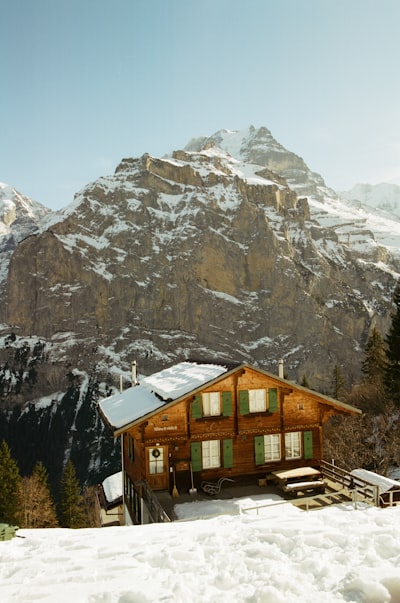 A Timber Mountain Hut With Green Shutters Set Against A Vast Snow-streaked Alpine Peak.