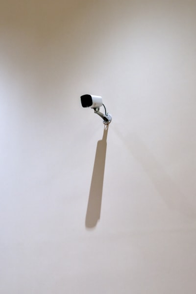 A Security Camera Mounted On A Blank Wall Casts A Long Shadow Under Soft Indoor Light. Surrounded By Empty Space, The Ordinary Object Becomes A Minimalist Study Of Surveillance, Geometry, And Presence.