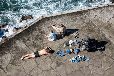 An Aerial View Of People Relaxing By The Ocean Near Funchal, Portugal. A Man Lies On The Ground, While Two Women Sit Together. Various Items Of Clothing And A Stroller Are Scattered Around Them.