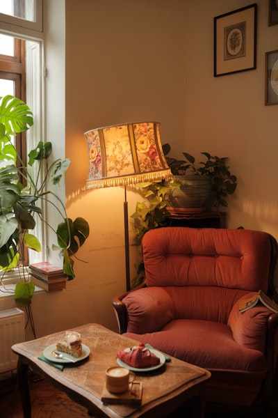 A Warm Café Corner With A Vintage Armchair, Floral Lampshade, Indoor Plants, Pastries, And Coffee On A Wooden Table, Creating A Nostalgic And Intimate Atmosphere. wallpaper preview