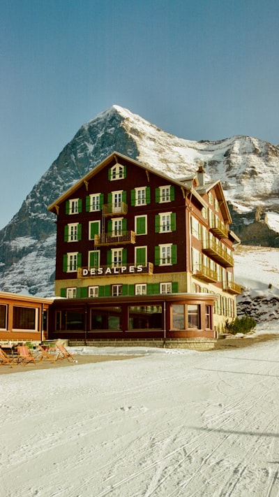 A Historic Chalet Hotel With Green Shutters Sits Below A Snowy Mountain Peak Under Clear Blue Sky. wallpaper preview