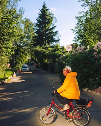 Older Person In Yellow Jacket Rides Red Bicycle On Street.