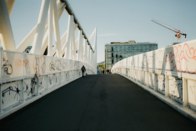 Person Walks On A White Pedestrian Bridge Covered In Graffiti.