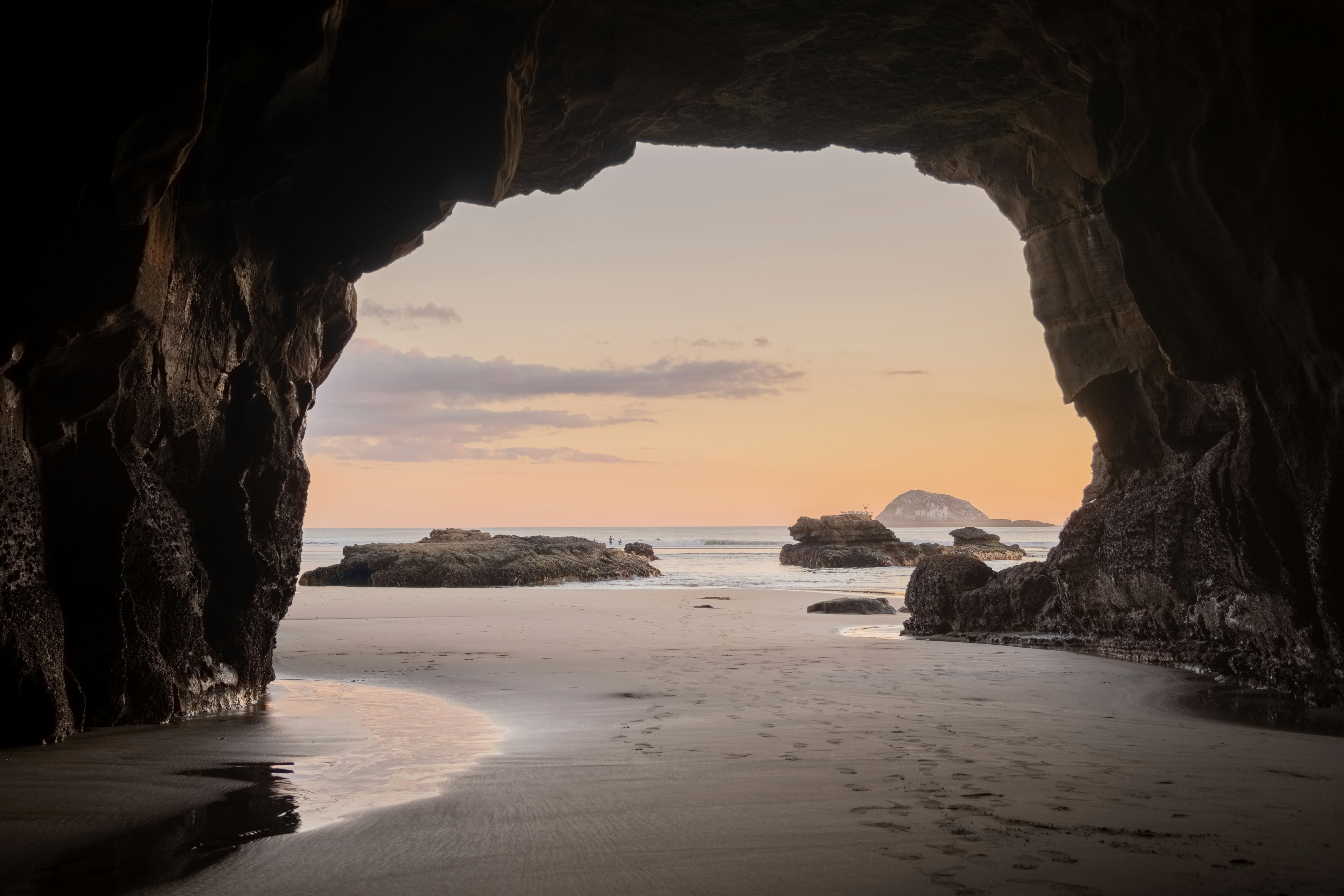 A Sea Cave Frames A Peaceful Beach Scene At Sunset, Drawing The Viewer's Eye Toward The Ocean And Distant Rock Formations. The Contrast Between The Dark Cave Interior And The Soft Pastel Sky Creates Depth And A Sense Of Exploration. HD mobile wallpaper