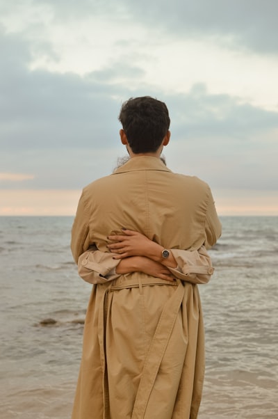 Person In Trench Coat Embraced By Another By The Ocean.