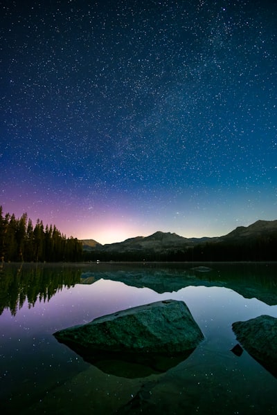 Starry Night Sky Over A Serene Lake With Mountain Reflections.
