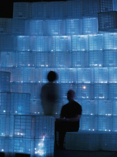 Two Figures Sit By A Glowing Blue Crate Wall.