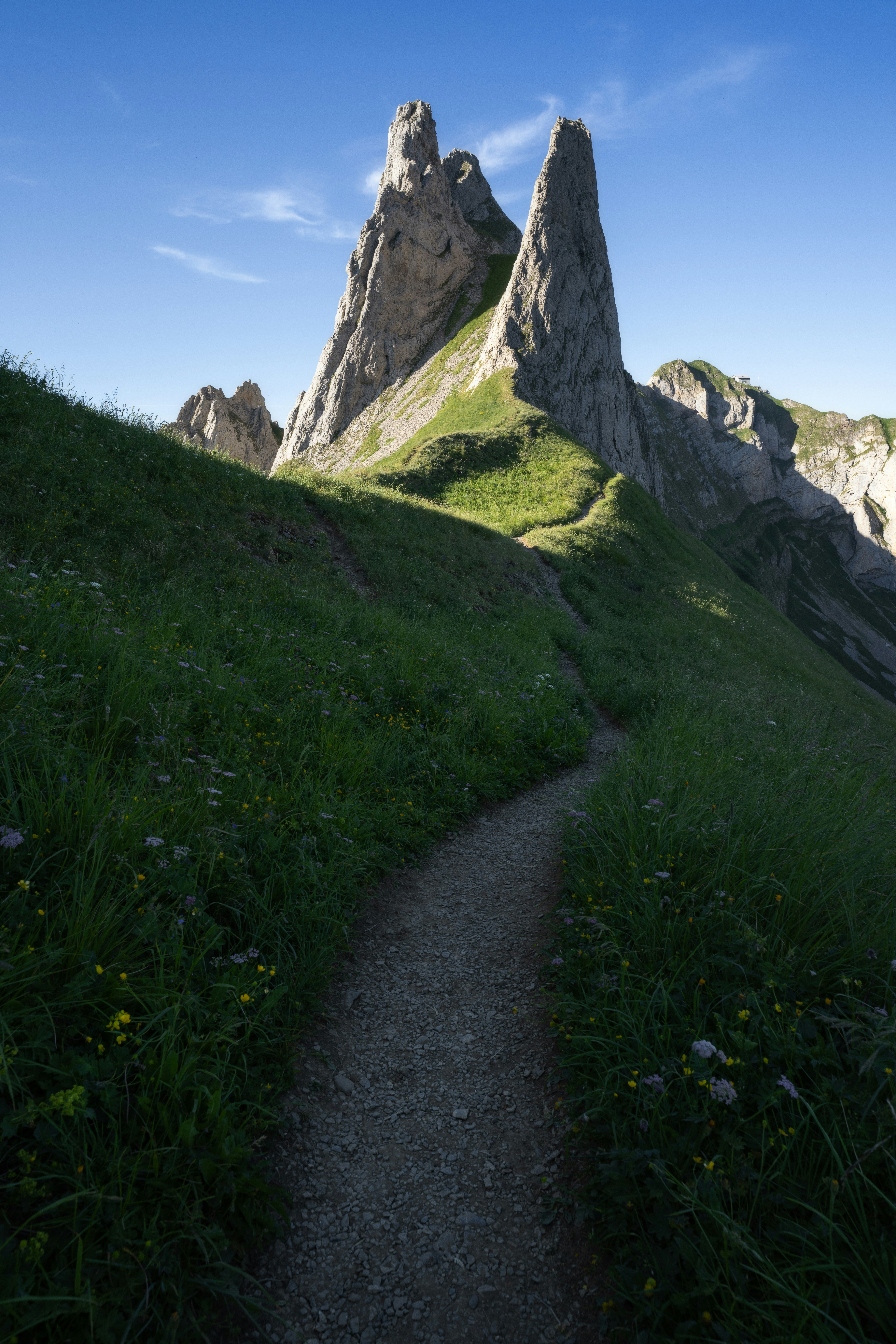 A Winding Mountain Trail Leads Toward Towering Limestone Peaks, Creating A Powerful Sense Of Depth And Adventure. Bathed In The Soft Light Of Early Morning, The Rugged Cliffs Rise Dramatically Against A Clear Blue Sky, While Lush Alpine Meadows Frame The Path In Vibrant Shades Of Green. The Scene Captures The Timeless Beauty Of The Mountains, Inviting Every Hiker To Explore The Wilderness One Step At A Time. HD mobile wallpaper